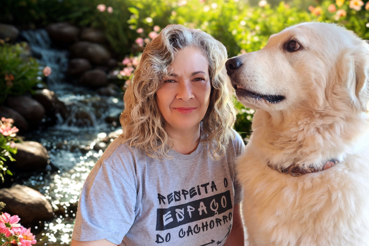 Foto minha com o Jack, meu kuvasz. Sou uma mulher branca, de meia idade, nessa foto estou com os cabelos um pouco abaixo dos ombros, com mechas loiras, ondulados. Uso uma blusa cinza e estou sentada ao lado do Jack, um cão de pelo branco longo. Atrás de nós tem uma paisagem feita por IA de um riacho e flores