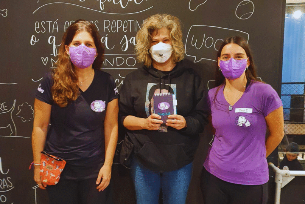 Foto minha, mulher branca, de meia idade, cabelos na altura dos ombros meio cacheados com mechas loiras. Estou entra as duas Carolinas da Turma do Focinho, ambas mulheres brancas, sendo a Carol Jardim à direita de cabelos longos loiros e à esquerda a Carol com cabelos escuros. Usamos máscaras no rosto por conta da pandemia.